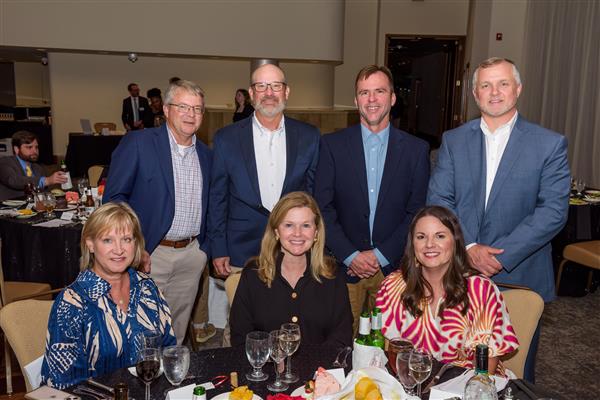 A group of seven well-dressed adults pose for a photo at a formal dinner event. Four individuals are standing behind a round table while three others are seated at the table. The table is set with glasses, plates of cake, salads, rolls, and bottles of wine and water. The background shows a large roo
